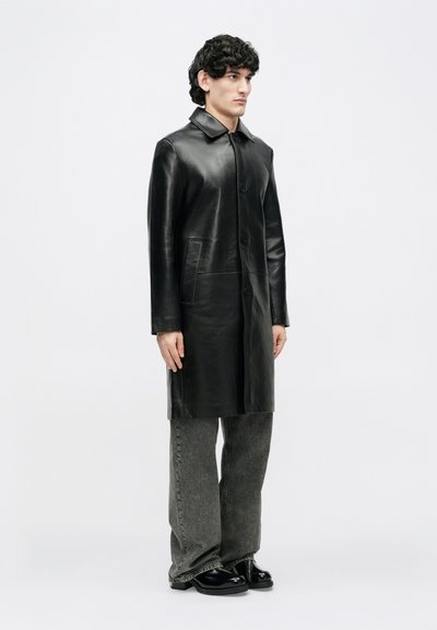 Abrigo de cuero negro con cuello, botones delanteros y dos bolsillos laterales. Llevado sobre pantalones grises de pierna ancha y zapatos negros. Textura lisa.