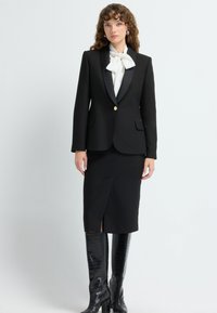 Žena v černém blejzru se saténovými klopami, bílou halenkou s mašlí, černé tužkové sukni s rozparkem a černých texturovaných nad kolena vysokých botách, stojící.