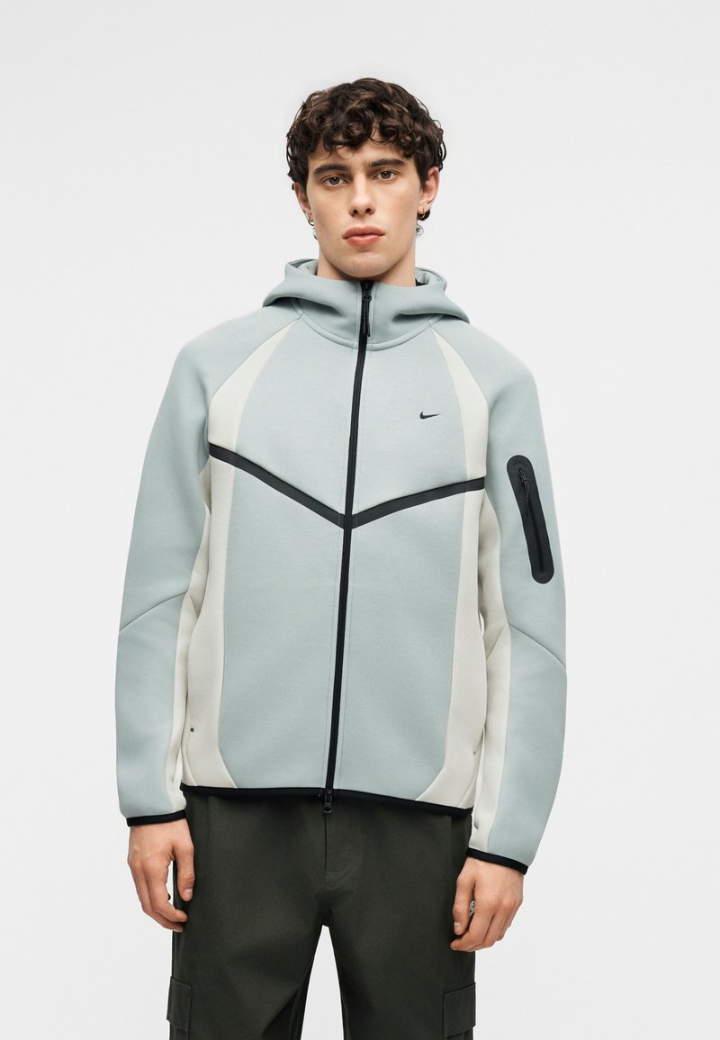 Junger Mann trägt einen hellgrau-weißen Kapuzenpullover mit Reißverschluss, schwarzen Reißverschlussdetails und Seitentasche, kombiniert mit dunklen Hosen.