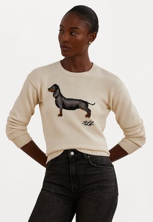 Pull en tricot crème avec un motif de teckel noir et des initiales brodées. Coupe ajustée avec des poignets et un col côtelés. Jeans foncés portés.