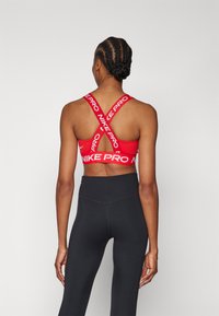 Sujeción deportiva roja Nike Pro con tirantes cruzados, con logotipo en blanco. Combinada con leggings negros de cintura alta. Textura de tela suave.