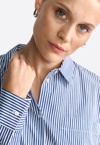 Blue and white striped button-up shirt with a collar, single chest pocket, and buttoned cuff. Smooth fabric texture. Silver earrings visible.