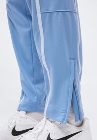 Ανοιχτό μπλε παντελόνια φόρμας με λευκές πλαϊνές ρίγες και φερμουάρ αστραγάλου φορεμένα πάνω από λευκά παπούτσια Nike με μαύρα λογότυπα swoosh.