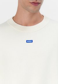 Kreemivalge kootud särk, millel on soonikuga kaelus ja sinine ristkülikukujuline "HUGO" logoplaat rinnal. Pehme tekstuur.