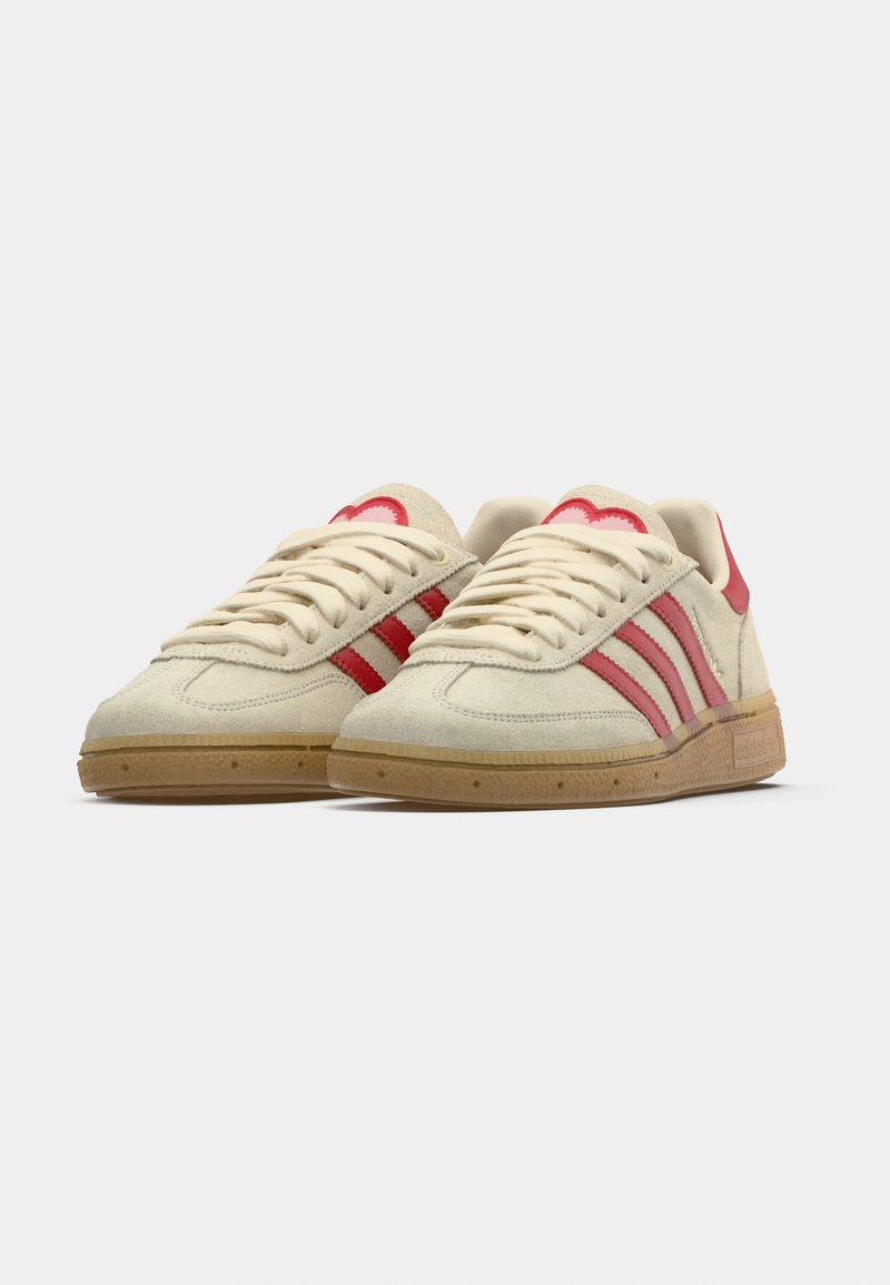 Light beige sneakers featuring red stripes and accents, suede material, round toe, and rubber gum sole. Laces are off-white.