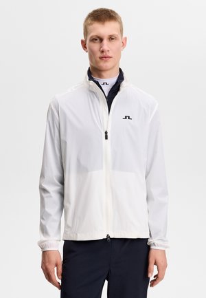 Jonge man met een wit jack met rits over een zwarte trui met hoge hals, tegen een effen lichte achtergrond.