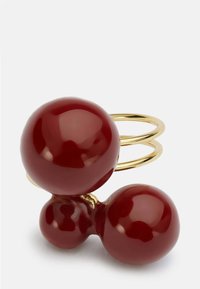 Rdeč sijajen izstopajoč prstan z značilnimi sferičnimi komponentami. Dve veliki rdeči krogli z bleščečo površino, povezani z zlatom obarvano kovinsko povezavo.