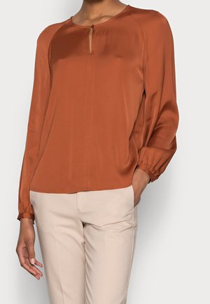 Femme portant un chemisier manches longues couleur rouille avec un petit bouton à l'avant et un pantalon beige, debout avec une main dans la poche.