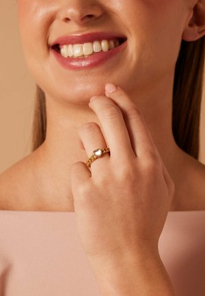 Femme souriante avec un rouge à lèvres rose touchant son menton, portant une bague en chaîne dorée avec un détail en forme de cœur blanc sur son doigt.