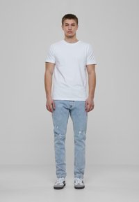 Camiseta blanca de algodón, jeans desgastados de color azul claro, zapatillas gris y blancas, fondo plano, figura masculina de pie.