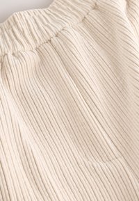 Béžová kordurová tkanina s vertikálnym rebrovaním a zbieraným pásom. Má jemné vrecko a mäkký, textúrovaný povrch.