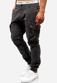Cargo byxor i mörkgrått tyg med flera fickor, en smal passform, ribbade muddar och en Calvin Klein-logotyp på midjan. Bärs med vita sneakers.