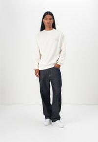 Cremefarbener strukturierter Sweatshirt mit gerippten Bündchen, kombiniert mit dunkelblauen Jeans und weißen Sneakers, vor einem schlichten weißen Hintergrund stehend.
