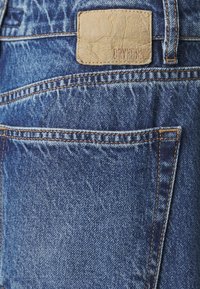 Modré džínsy z denimu s textúrovaným povrchom, s hnedou koženou nášivkou a oranžovým prešívaním pozdĺž vreciek a švov.