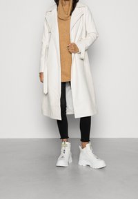 Cappotto lungo bianco, tessuto strutturato, vita con cintura, indossato sopra un maglione marrone lavorato a maglia, abbinato a jeans neri e stivali bianchi robusti.