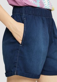 Shorts en denim avec un waistband élastique, couleur bleu foncé, poches latérales et détails de ourlet roulé. Texture lisse avec accents cousus.