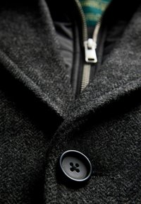 Sort uldblandingfrakke med en markant knap, tekstureret stof og en lynlåslukning med en kontrasterende grøn foring, der er synlig indeni.