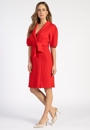 Red wrap dress with short, puffed sleeves, a tie waist, and knee-length hem. Worn with nude high-heeled shoes on a neutral background.