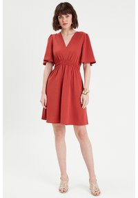 Vestido color óxido con un escote en V profundo, mangas cortas con vuelo, cintura fruncida y una falda fluida, combinado con sandalias plateadas de tiras.