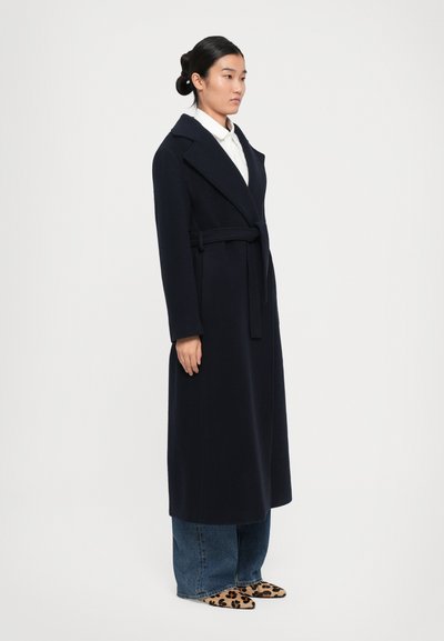 Donna che indossa un lungo cappotto blu con cintura sopra un paio di jeans blu e scarpe con stampa leopardata, in piedi contro uno sfondo chiaro e neutro.