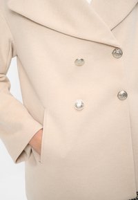 Dubbelknäppt beige kappa med en texturerad tyg. Har stora silverknappar och sidofickor. Breda slag och avslappnad passform.