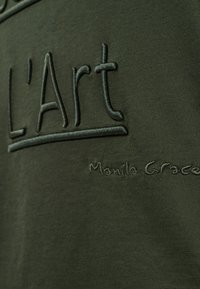Tissu en coton vert olive avec un texte brodé "L'Art" et "Manila Grace" dans un point contrasté, présentant une texture douce et un design minimaliste.