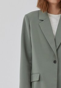Lysegrønn blazer i glatt stoff med notched krage, enkel svart knapp og to frontlommer. Brukt over en lys grå topp.