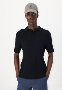 Mörkblå, stickad polo shirt med korta ärmar, som har en strukturerad flätad design och en klassisk krage; ihop med en ljusgrå keps.