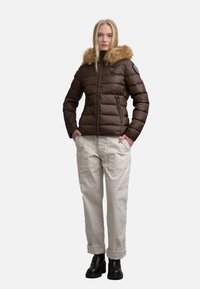 Giacca imbottita marrone con cappuccio foderato in pelliccia, caratterizzata da una chiusura frontale con zip e un design trapuntato. Abbinate a pantaloni chiari e stivali neri.