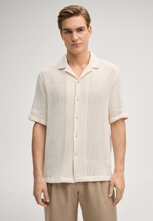 Giovane uomo che indossa una maglia di maglia color crema con maniche corte e bottoni, abbinata a pantaloni beige, su uno sfondo chiaro e uniforme.