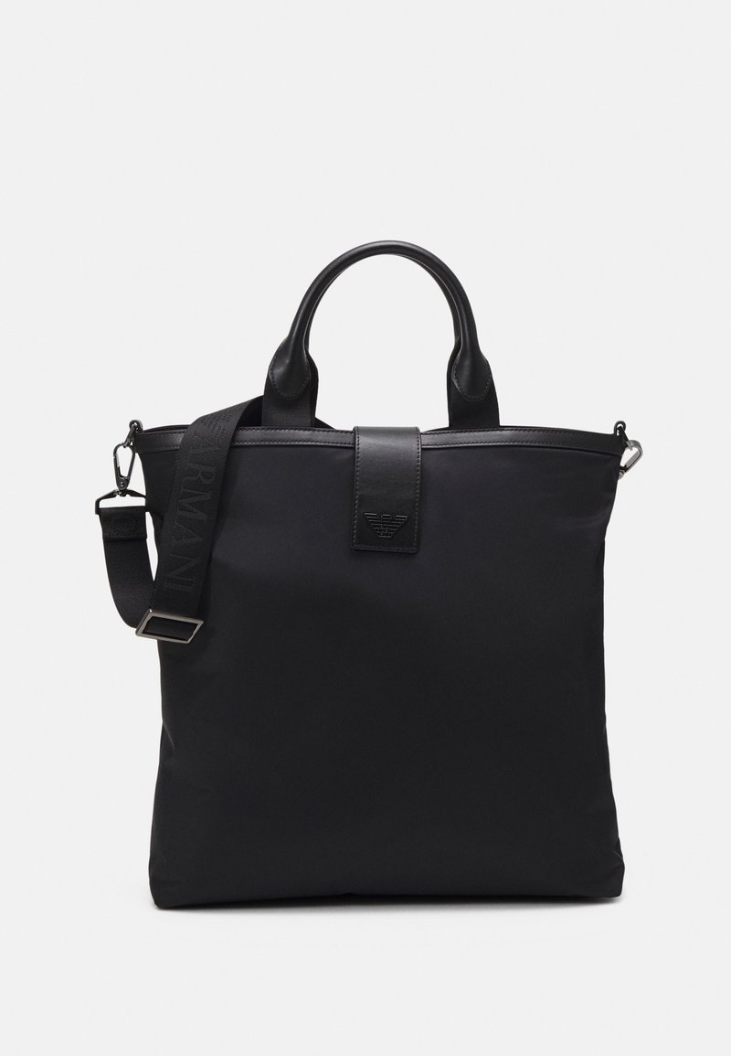 Sac cabas noir avec poignées en cuir, bandoulière amovible et détail du logo sur le rabat avant.