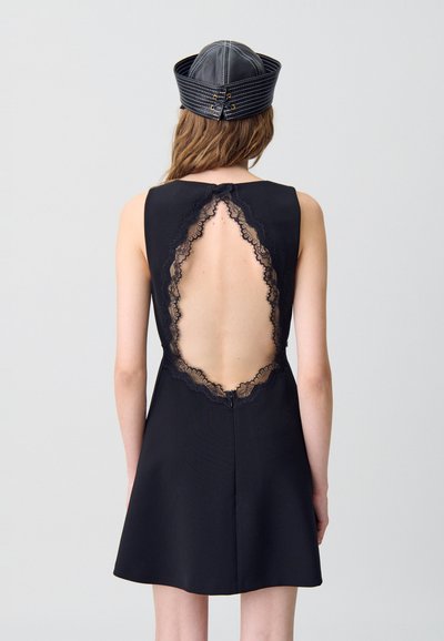 Donna che indossa un vestito nero senza maniche con una grande schiena scoperta bordata di pizzo e un cappello nero di pelle cucito, in piedi davanti a uno sfondo chiaro e semplice.