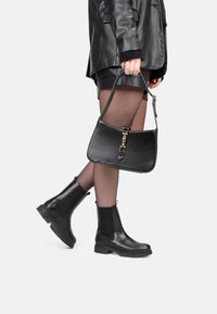 Bolso de cuero negro con herrajes dorados y un acento en forma de lágrima. Botas de tobillo negras con acabado texturizado y paneles laterales elásticos.
