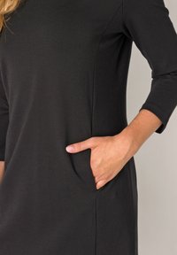 Zwarte jurk met driekwart mouwen, voorzien van een zijzak en een gladde, rekbare stof. Eenvoudige silhouet met strakke lijnen, zonder patronen.