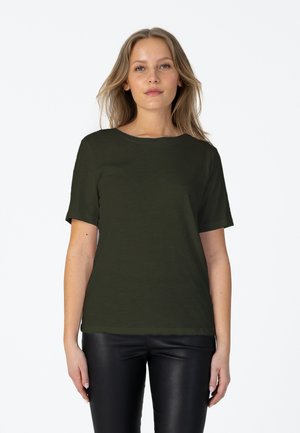 Femme aux longs cheveux blonds portant une chemise à manches courtes vert foncé et un pantalon en cuir noir, debout sur un fond blanc.