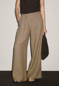 Braune, weit geschnittene Hose aus strukturiertem Stoff mit hoher Taille und Falten, kombiniert mit einer schwarzen Tasche und hellen Sandalen.