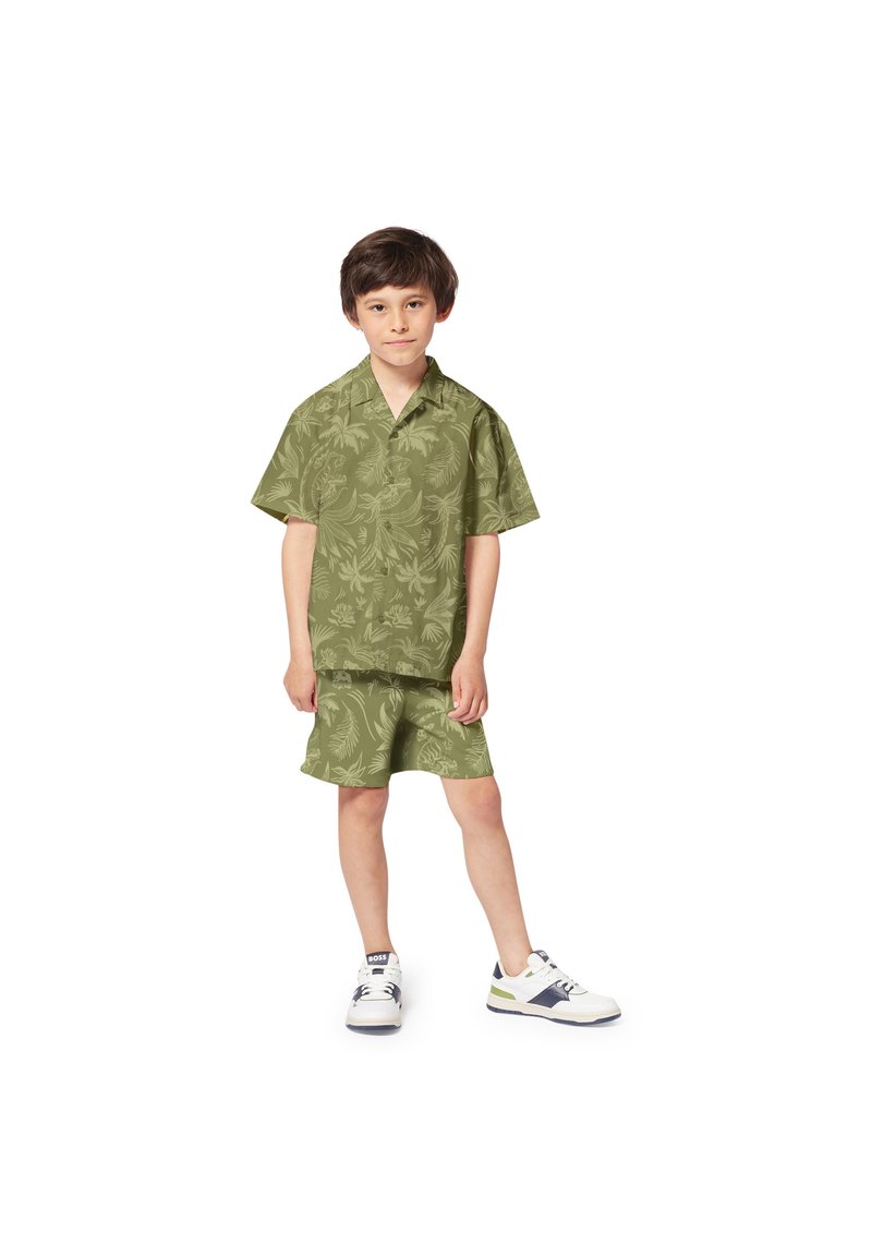 Olijf groene shirt met korte mouwen en een bladpatroon, gecombineerd met bijpassende shorts. Heeft een relaxte pasvorm. Gedragen met witte sneakers.