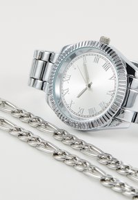 Montre analogique argentée avec une lunette striée, des marqueurs en chiffres romains et un bracelet en acier inoxydable poli, accompagnée d'une chaîne argentée assortie.