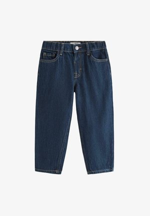 Smilende ung dreng står med den ene hånd i siden iført en enkel hvid T-shirt, mørkeblå jeans og hvide sneakers mod en ensfarvet baggrund.