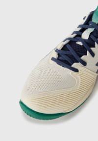 White and light cream athletic shoe with textured upper, navy laces, green outsole, and a ridged toe box.