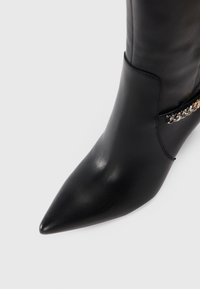 Black leather ankle boot with a pointed toe, smooth texture, and a decorative gold chain accent on the side.