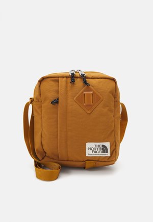 Kompakte senffarbene Schultertasche mit Frontreißverschlusstasche, verstellbarem Trageriemen und „The North Face“-Logoaufnäher unten rechts.