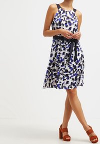 Robe à fleurs avec une base blanche ornée de motifs bleus et noirs, un corsage ajusté et une ceinture à nouer noire à la taille. Sandales à talons blocs marron.