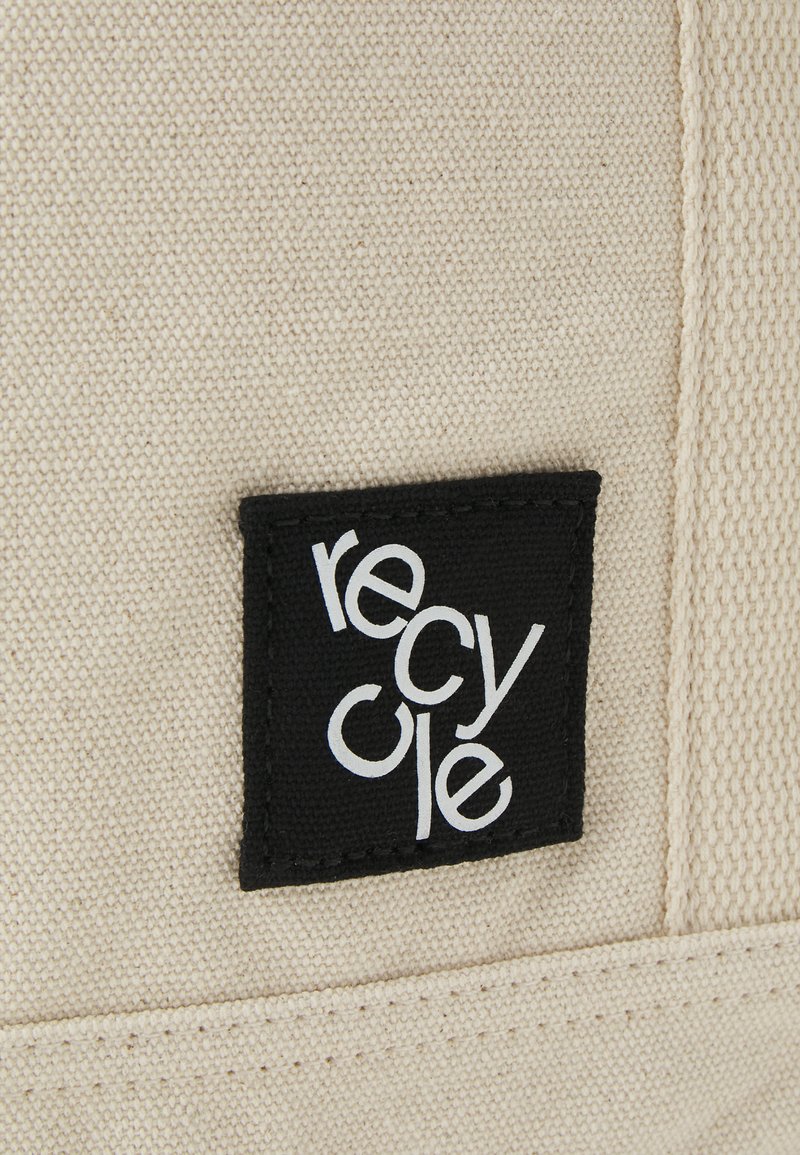 Detalle de una bolsa de lona que muestra una etiqueta negra con el texto "reciclar" en blanco. La bolsa es de color crema y tiene una superficie texturizada.