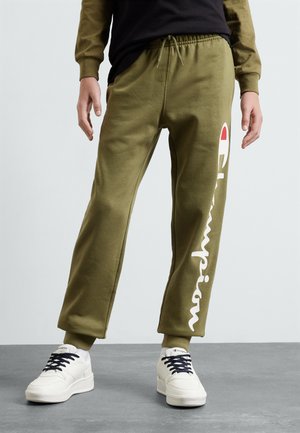 Olijfgroene joggingbroek met een elastische tailleband, zijzakken en een witte "Champion"-logo dat verticaal op het linkerbeen is gedrukt, gecombineerd met witte sneakers.