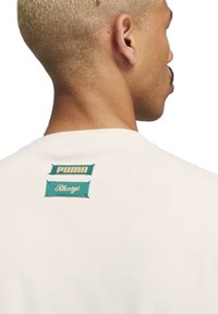 Witte T-shirt met twee gestikte labels op de bovenrug: een groen rechthoekig label met "PUMA" in goud en een ander label met "Rhuigi" in witte script.