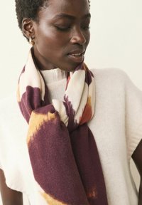 Strikket tørklæde med en cremet bund og lilla og rustfarvede tie-dye mønstre. Blød tekstur, draperet omkring halsen, parret med en lys sweater.