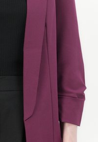 Bordeauxblazer med öppen front, strukturerade axlar och långa ärmar, tillsammans med en svart ribbad topp och högmidjade mörka byxor.