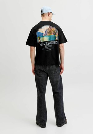 Uomo che indossa una maglietta nera con stampa grafica colorata di un villaggio turistico sul lago e jeans neri, di spalle, su sfondo grigio uniforme.
