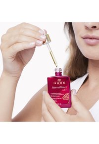 Rosa Glasflasche des NUXE Merveillance LIFT Serums mit einem goldenen Tropfenverschluss. Klare Flüssigkeit wird abgegeben. Etikett sichtbar mit Produktdetails.